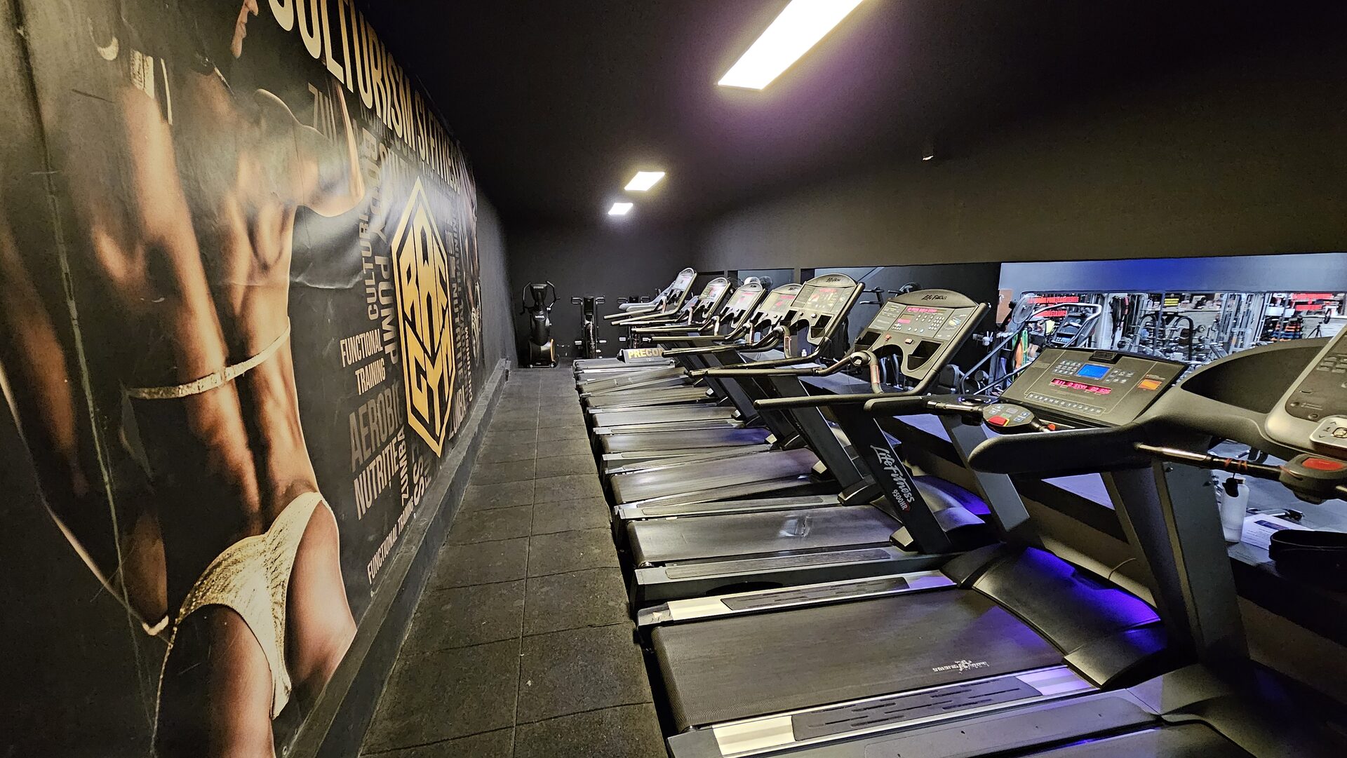 Zona de cardio, goală: opt benzi de alergare aliniate de-a lungul peretelui pe care e pictat „CULTURISM ȘI FITNESS".